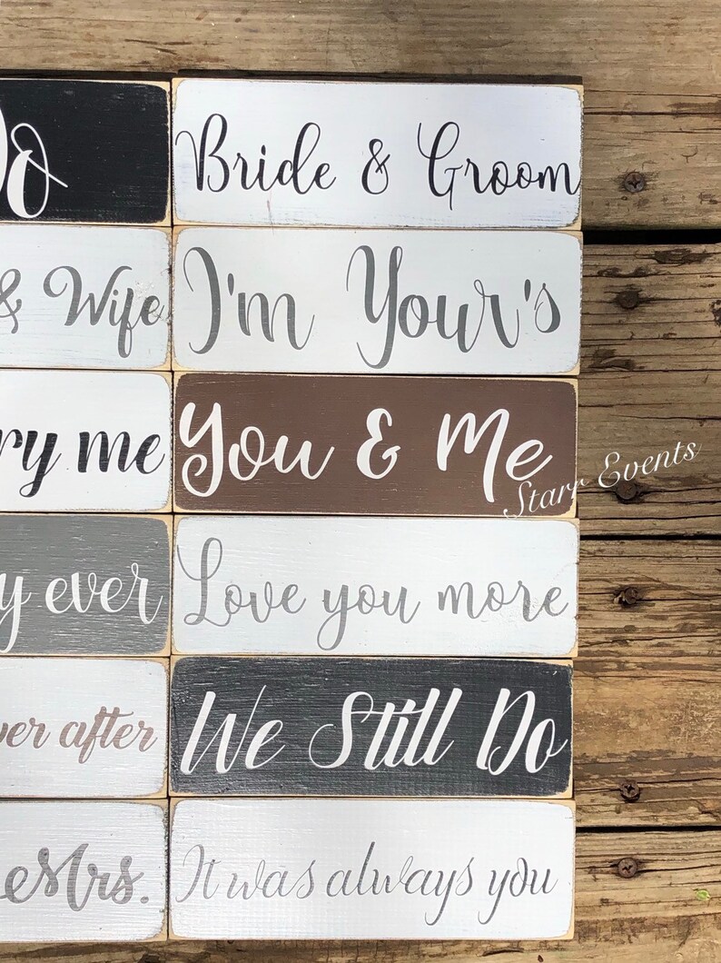 May include: A collection of rectangular wooden signs with various wedding-themed phrases in elegant script. The signs are painted in a variety of colors, including black, white, gray, and brown. Phrases include "Bride & Groom" and "We Still Do."