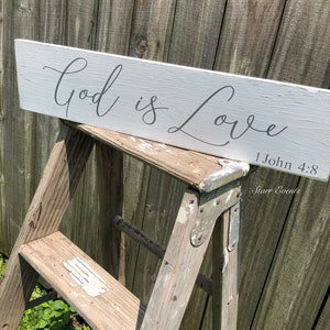 May include: A white wooden sign with the text "God is Love" in gray script. The sign also includes the Bible verse 1 John 4:8. The sign is displayed on a wooden ladder.