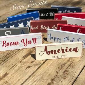 May include: A selection of rectangular wooden signs with patriotic themes. The signs are painted in red, white, and blue, with various phrases and the word "America" displayed. The signs are arranged on a wooden surface.