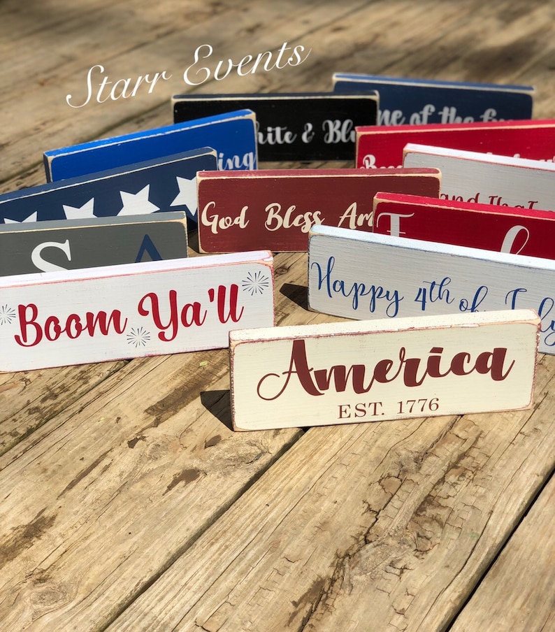May include: A selection of rectangular wooden signs with patriotic themes. The signs are painted in red, white, and blue, with various phrases and the word "America" displayed. The signs are arranged on a wooden surface.