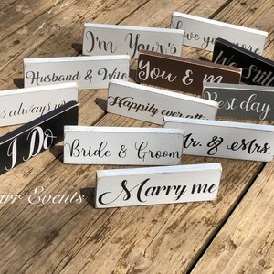 May include: A collection of small, rectangular wooden signs with various wedding-themed phrases in elegant script. The signs are painted in neutral colors like white, gray, and black, with phrases such as "I Do" and "Marry Me".