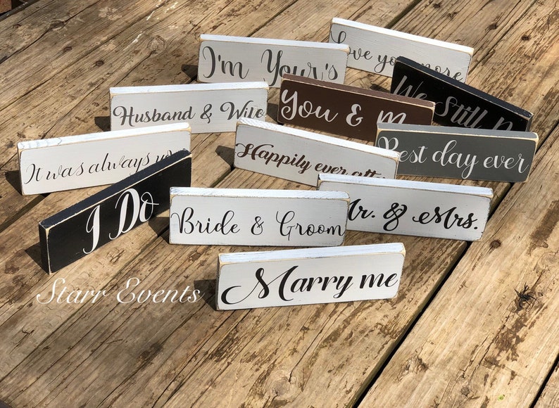 May include: A collection of small, rectangular wooden signs with various wedding-themed phrases in elegant script. The signs are painted in neutral colors like white, gray, and black, with phrases such as "I Do" and "Marry Me".