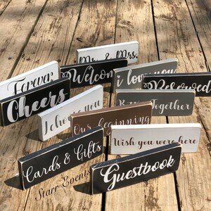 May include: A selection of small, rectangular wooden signs with white lettering on black, white, grey, and brown backgrounds. The signs display phrases such as "Forever," "Welcome," "I love you," and "Guestbook." The signs are arranged on a weathered wooden surface.