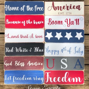 May include: A selection of rectangular wooden signs with patriotic slogans in red, white, and blue. Phrases include "Home of the free," "America," "Because of the brave," and "Happy 4th of July." The signs are displayed on a wooden surface.