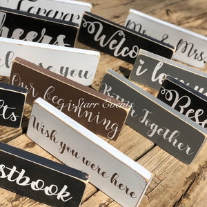 May include: A selection of rectangular wooden signs, each bearing a cursive phrase. The signs are painted in white, black, grey, and brown. Phrases include "Welcome," "Forever," and "Wish you were here."