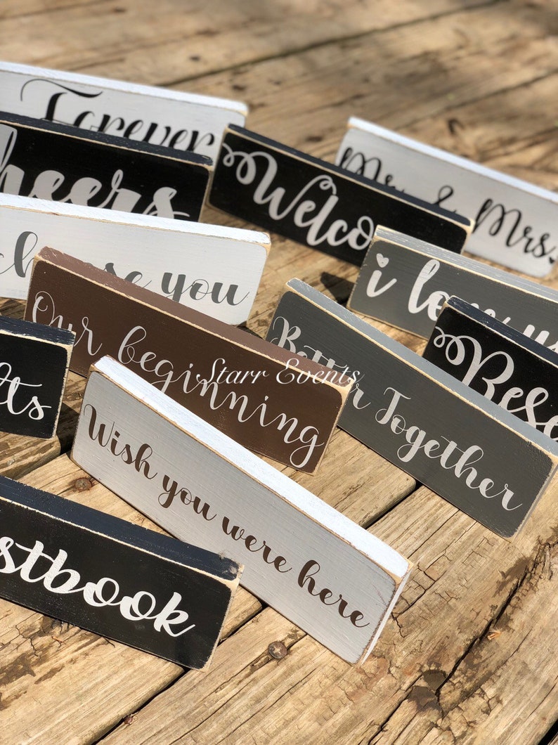 May include: A selection of rectangular wooden signs, each bearing a cursive phrase. The signs are painted in white, black, grey, and brown. Phrases include "Welcome," "Forever," and "Wish you were here."