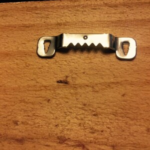 May include: Close-up of a wooden surface with a metal sawtooth picture hanger attached. The hanger is silver and has two keyhole slots on either end and a series of teeth in the middle for secure hanging.