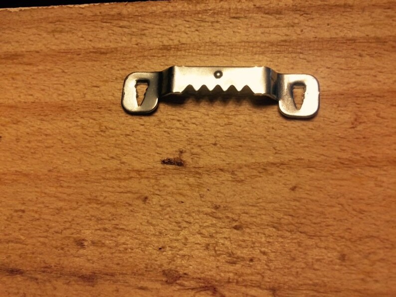 May include: Close-up of a wooden surface with a metal sawtooth picture hanger attached. The hanger is silver and has two keyhole slots on either end and a series of teeth in the middle for secure hanging.