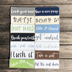 May include: A collection of rectangular wooden signs with various bathroom-related phrases in white script. The signs are painted in different colors, including gray, navy, beige, green, black, blue, white, brown, and light green. The phrases include "Comb your hair," "Have a nice poop," and "Flush it."