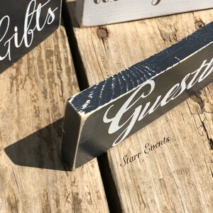 May include: Close-up of wooden signs with white script on a distressed black and grey background. The signs read "Gifts", "Wish you were", and "Guests". The signs are on a rustic wooden surface.