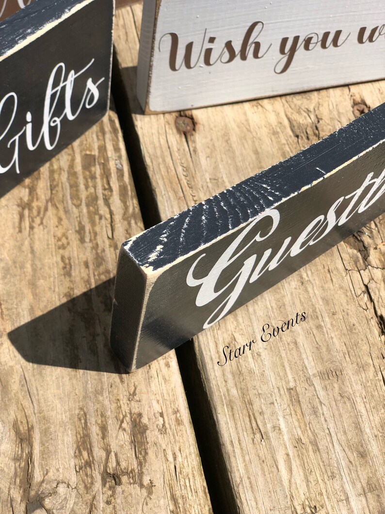 May include: Close-up of wooden signs with white script on a distressed black and grey background. The signs read "Gifts", "Wish you were", and "Guests". The signs are on a rustic wooden surface.