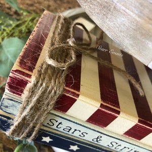 Puede incluir: Primer plano de una pieza decorativa de madera con tem&aacute;tica patri&oacute;tica. Presenta rayas rojas y blancas, con las palabras "Stars & Stripes" visibles. La pieza est&aacute; atada con cuerda y tiene un acabado r&uacute;stico y envejecido.