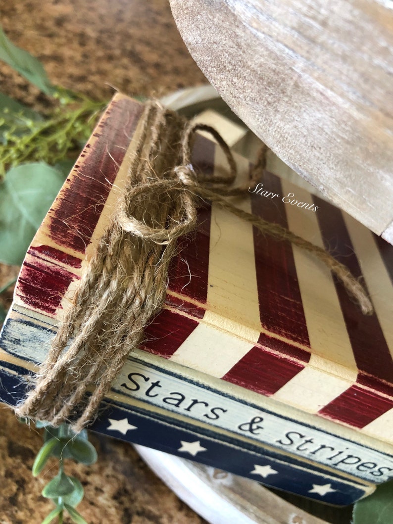 Puede incluir: Primer plano de una pieza decorativa de madera con tem&aacute;tica patri&oacute;tica. Presenta rayas rojas y blancas, con las palabras "Stars & Stripes" visibles. La pieza est&aacute; atada con cuerda y tiene un acabado r&uacute;stico y envejecido.