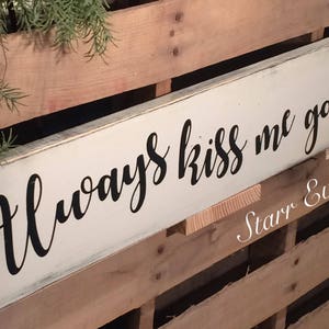 May include: A rectangular, cream-coloured wooden sign with the words "Always kiss me goodnight" in black script. The sign is mounted on a wooden pallet background, with greenery and burlap accents.