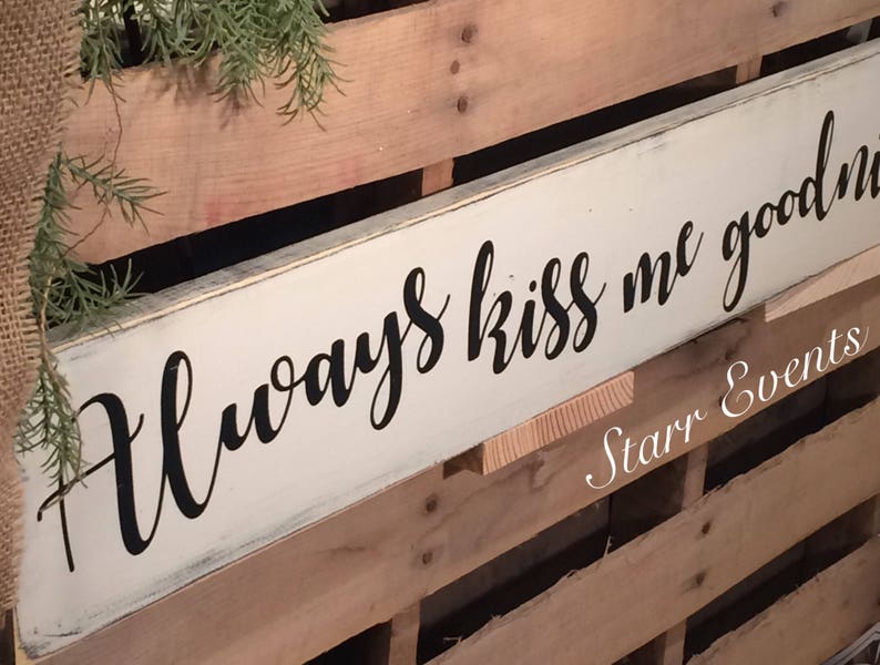 May include: A rectangular, cream-coloured wooden sign with the words "Always kiss me goodnight" in black script. The sign is mounted on a wooden pallet background, with greenery and burlap accents.
