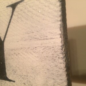 May include: Close-up of a distressed wooden sign with the letter "Y" in black against a white background. The wood has a textured, weathered appearance, with visible grain and a slightly rough edge. The sign is likely for home decor.