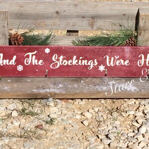 Pode incluir: Uma placa retangular de madeira bord&ocirc; com a frase "And The Stockings Were Hung" em escrita branca. A placa tem tr&ecirc;s ganchos e detalhes de flocos de neve. Decorada com pinhas, vegeta&ccedil;&atilde;o e bagas vermelhas.
