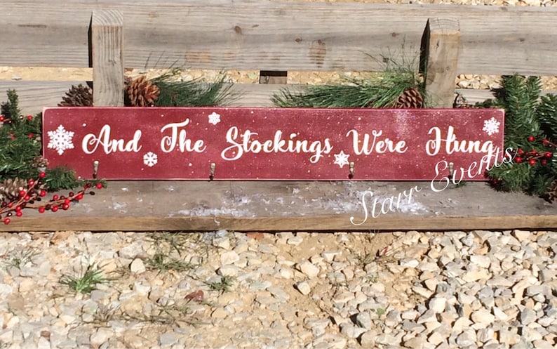 Pode incluir: Uma placa retangular de madeira bord&ocirc; com a frase "And The Stockings Were Hung" em escrita branca. A placa tem tr&ecirc;s ganchos e detalhes de flocos de neve. Decorada com pinhas, vegeta&ccedil;&atilde;o e bagas vermelhas.