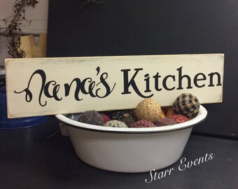 May include: A cream-colored wooden sign with the words "Nana's Kitchen" in black script. The sign rests above a white enamel bowl filled with fabric-covered decorative balls in various patterns and colors. A charming kitchen decor item.