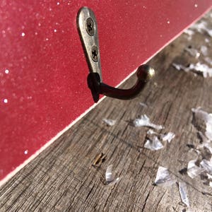 Pode incluir: Close-up de um gancho de parede decorativo. O gancho &eacute; feito de metal escuro e &eacute; fixado a uma superf&iacute;cie vermelha e brilhante com dois parafusos. O gancho &eacute; montado em uma superf&iacute;cie de madeira desgastada.