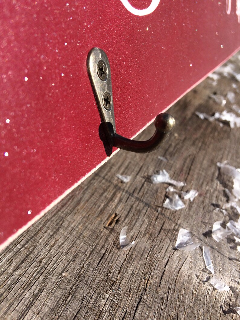 Pode incluir: Close-up de um gancho de parede decorativo. O gancho &eacute; feito de metal escuro e &eacute; fixado a uma superf&iacute;cie vermelha e brilhante com dois parafusos. O gancho &eacute; montado em uma superf&iacute;cie de madeira desgastada.