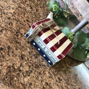 Puede incluir: Una pila de bloques de madera decorativos atados con hilo. Los bloques est&aacute;n pintados con un tema patri&oacute;tico, con rayas rojas y blancas y una secci&oacute;n azul con estrellas blancas. Las palabras "Stars & Stripes" son visibles.