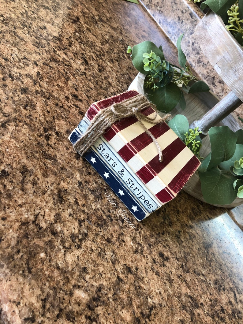 Puede incluir: Una pila de bloques de madera decorativos atados con hilo. Los bloques est&aacute;n pintados con un tema patri&oacute;tico, con rayas rojas y blancas y una secci&oacute;n azul con estrellas blancas. Las palabras "Stars & Stripes" son visibles.