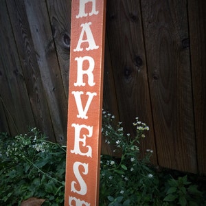 May include: An orange wooden sign with the word "HARVEST" in white lettering. The sign is placed vertically against a wooden fence, with green plants and small white flowers in the foreground. A decorative item for the home.