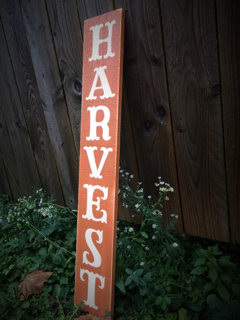 May include: An orange wooden sign with the word "HARVEST" in white lettering. The sign is placed vertically against a wooden fence, with green plants and small white flowers in the foreground. A decorative item for the home.