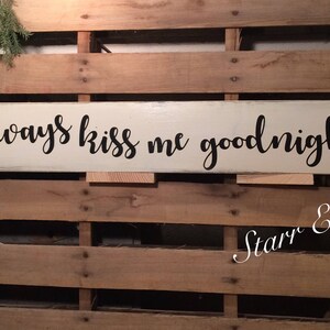 May include: An off-white wooden sign with the words "Always kiss me goodnight" in black script. The sign is set against a backdrop of wooden planks. A sprig of greenery is visible in the upper left corner.