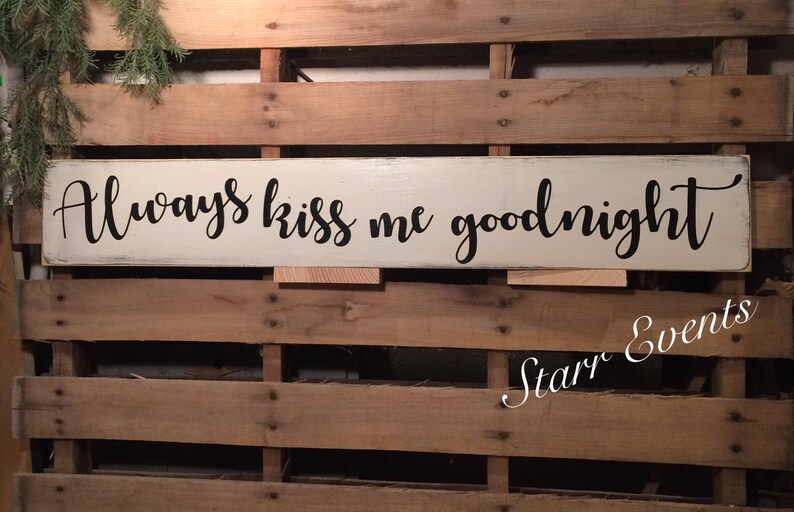 May include: An off-white wooden sign with the words "Always kiss me goodnight" in black script. The sign is set against a backdrop of wooden planks. A sprig of greenery is visible in the upper left corner.