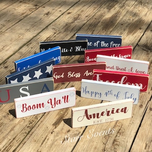 May include: A selection of rectangular wooden signs in various colours, including red, white, blue, and grey. Each sign features patriotic phrases such as "America," "Freedom," and "Happy 4th of July" in white or red lettering. The signs are arranged on a weathered wooden surface.