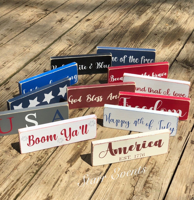 May include: A selection of rectangular wooden signs in various colours, including red, white, blue, and grey. Each sign features patriotic phrases such as "America," "Freedom," and "Happy 4th of July" in white or red lettering. The signs are arranged on a weathered wooden surface.