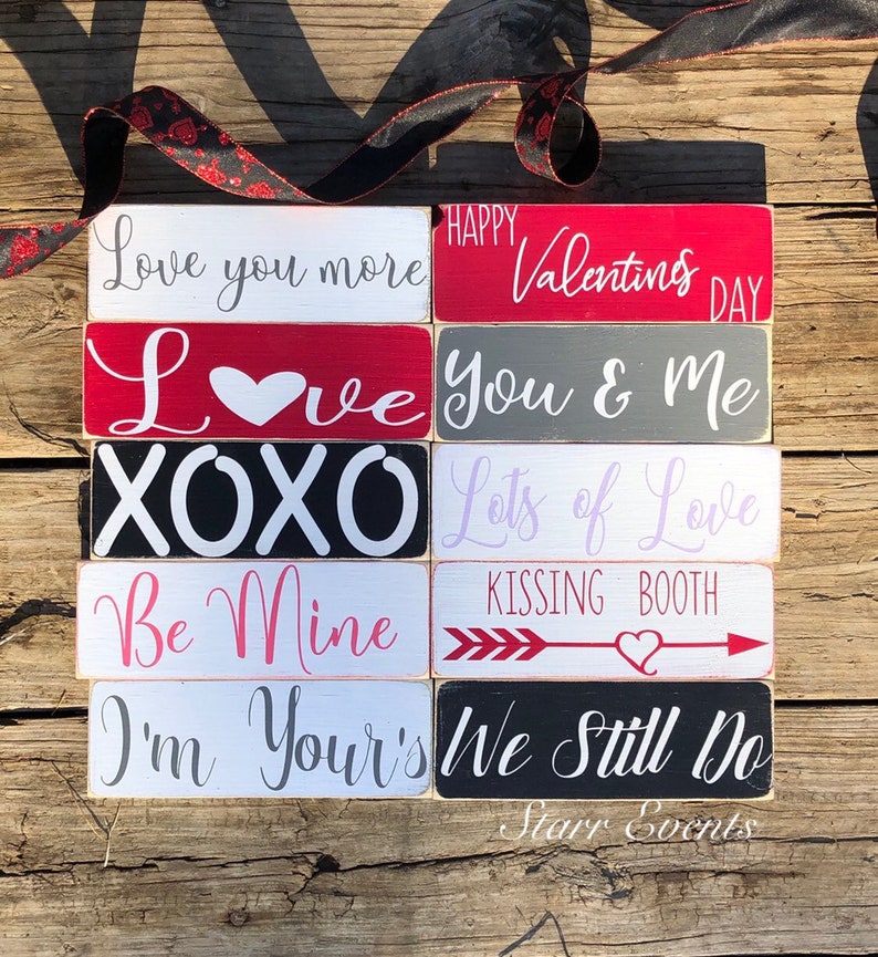 May include: Ten small Valentine's Day themed wooden blocks with various romantic phrases. The blocks feature phrases like 'Love you more', 'Happy Valentine's Day', 'XOXO', 'Lots of Love', 'Be Mine', 'I'm Yours', and 'We Still Do'. The blocks are painted in red, gray, black, white, and pink. These are perfect for Valentine's Day decor or gifts.