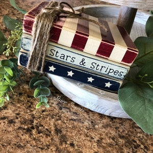 Puede incluir: Una pila de tres bloques de madera decorativos atados con cuerda. El bloque superior tiene rayas rojas y blancas, el bloque central es azul con estrellas blancas y el bloque inferior es blanco con rayas rojas. Las palabras "Stars & Stripes" est&aacute;n impresas.