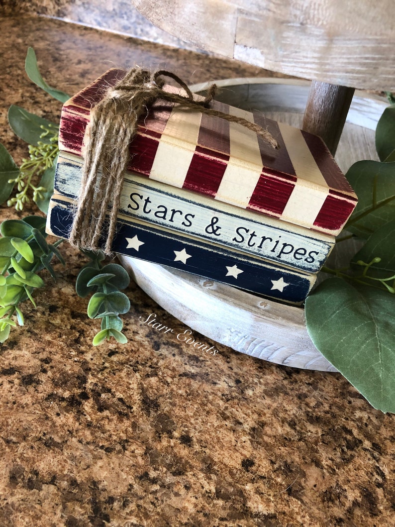 Puede incluir: Una pila de tres bloques de madera decorativos atados con cuerda. El bloque superior tiene rayas rojas y blancas, el bloque central es azul con estrellas blancas y el bloque inferior es blanco con rayas rojas. Las palabras "Stars & Stripes" est&aacute;n impresas.
