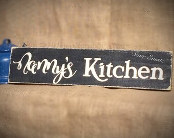 May include: A rectangular, distressed black wooden sign with the words "Nanny's Kitchen" in white script. The sign also includes the text "Starr Events" in a smaller font. A blue object is visible on the left side of the sign.