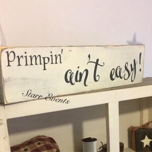May include: A distressed white wooden sign with the phrase "Primpin' ain't easy!" in black script. The sign also includes the text "Starr Events". A metal star decorates the shelf.