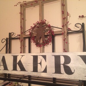 May include: A rustic bakery sign with the word "BAKERY" in large black letters on a distressed white wooden plank. The sign is mounted on a black metal frame, with a decorative star and berry wreath.