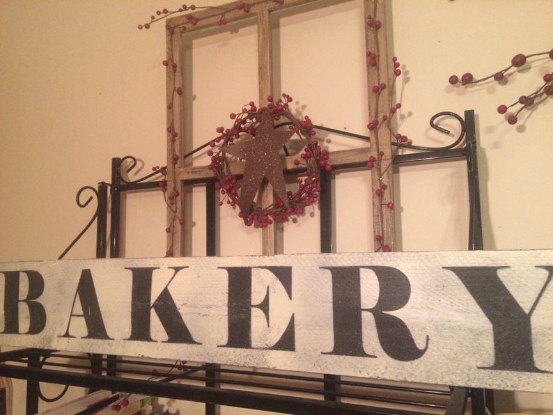 May include: A rustic bakery sign with the word "BAKERY" in large black letters on a distressed white wooden plank. The sign is mounted on a black metal frame, with a decorative star and berry wreath.