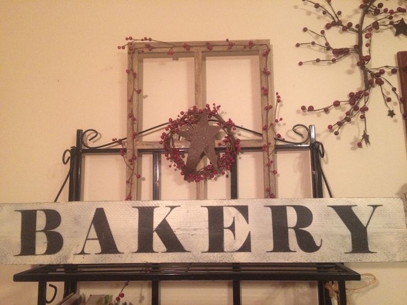 May include: A rustic wooden sign with the word "BAKERY" in large black letters. The sign is mounted on a black metal frame, with a decorative window frame and star-shaped wreath above it. Red berries and a star add to the country charm.