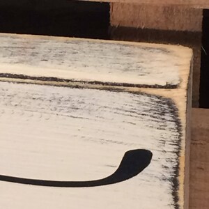 May include: Close-up of a distressed, white-painted wooden sign with a black, curved design element. The edges of the sign are dark, creating a rustic, aged appearance. The wood grain is visible, adding texture to the surface.