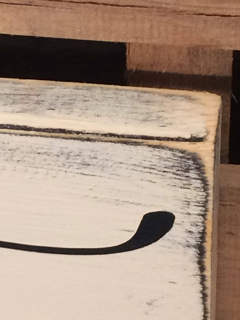 May include: Close-up of a distressed, white-painted wooden sign with a black, curved design element. The edges of the sign are dark, creating a rustic, aged appearance. The wood grain is visible, adding texture to the surface.