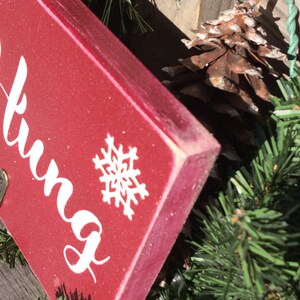 Pode incluir: Uma placa de madeira vermelha com escrita branca e um desenho de floco de neve. A placa faz parte de uma decora&ccedil;&atilde;o de Natal, rodeada por pinheiro verde, bagas vermelhas e uma pinha. As bordas da placa s&atilde;o ligeiramente desgastadas.