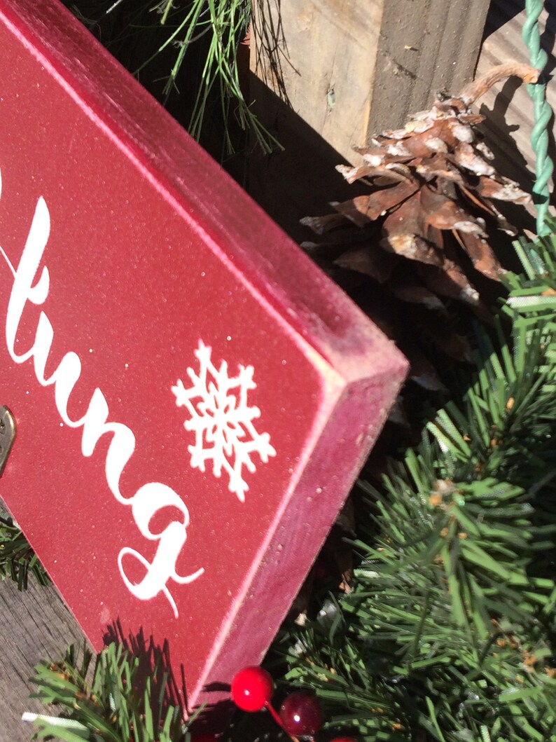 Pode incluir: Uma placa de madeira vermelha com escrita branca e um desenho de floco de neve. A placa faz parte de uma decora&ccedil;&atilde;o de Natal, rodeada por pinheiro verde, bagas vermelhas e uma pinha. As bordas da placa s&atilde;o ligeiramente desgastadas.