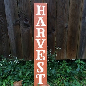 May include: A rectangular wooden sign, painted reddish-brown, with the word "HARVEST" in white, decorative lettering. The sign is placed in a garden, with a wooden fence behind it. The sign is approximately 120 cm tall.