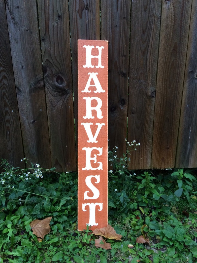May include: A rectangular wooden sign, painted reddish-brown, with the word "HARVEST" in white, decorative lettering. The sign is placed in a garden, with a wooden fence behind it. The sign is approximately 120 cm tall.