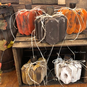 May include: A collection of rustic wooden pumpkins in various colors, including orange, brown, and white. Each pumpkin has a wooden stem and is adorned with raffia. The pumpkins are displayed on a wooden crate, creating a fall-themed display.
