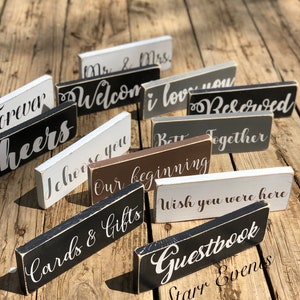 May include: A selection of rectangular wooden signs with white script phrases including "Welcome", "I love you", and "Cards & Gifts". The signs are painted in black, white, grey, and brown, and are arranged on a wooden surface.