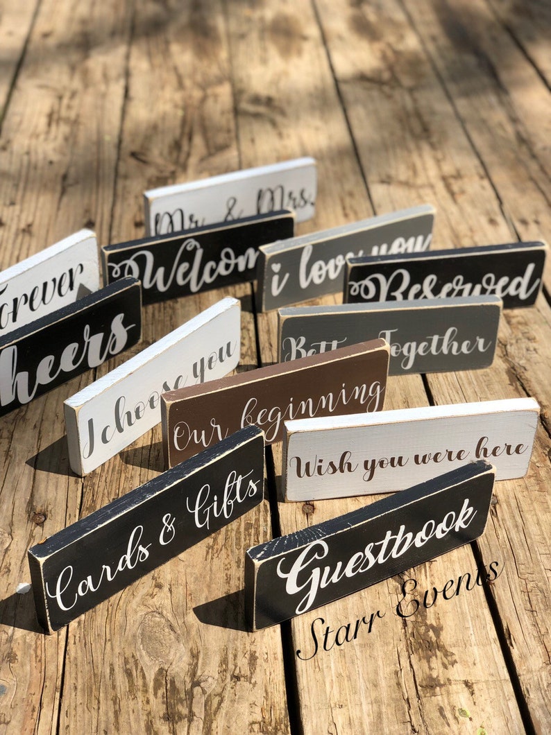 May include: A selection of rectangular wooden signs with white script phrases including "Welcome", "I love you", and "Cards & Gifts". The signs are painted in black, white, grey, and brown, and are arranged on a wooden surface.